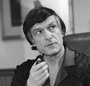 Black and white portrait of a young Hugh Hefner wearing his iconic silk pajama shirt, looking to the side while holding a smoking pipe to his chin.
